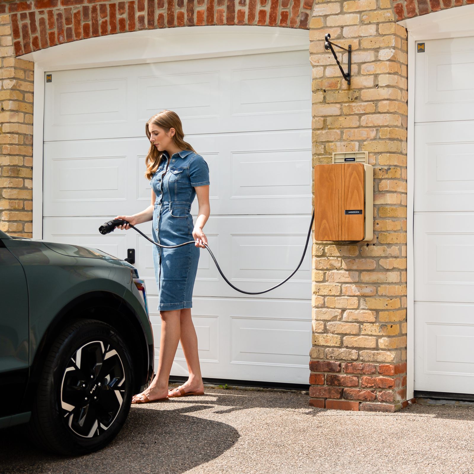 A homeowner connecting a cable from a wall-mounted home charger to an electric car in front of a garage